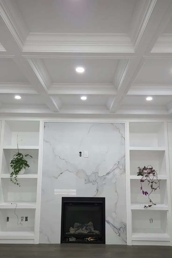 Coffered ceiling millwork above a veined panel fireplace wall with white built-in shelving